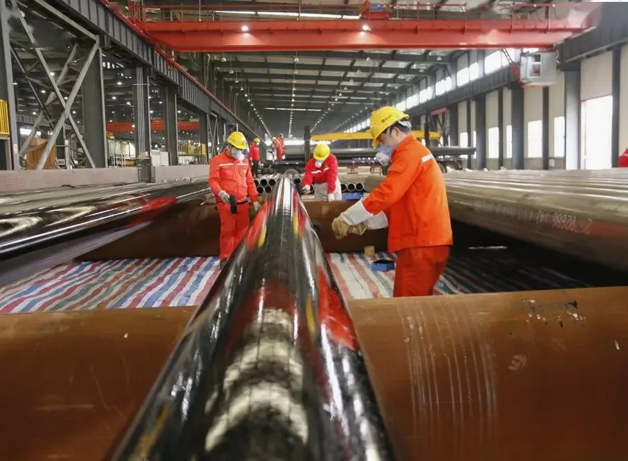 Workers handling long steel pipes with overhead crane slings in a heavy industrial fabrication workshop